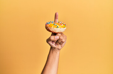 Finger of hispanic man holding donut over isolated yellow background.