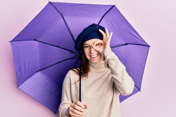 Young red head girl holding purple umbrella wearing fresh beret smiling happy doing ok sign with...
