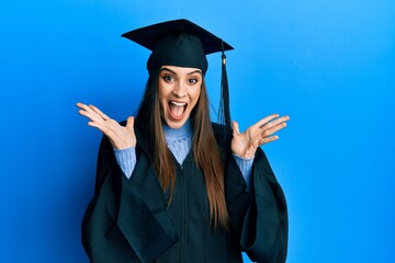 Beautiful brunette young woman wearing graduation cap and ceremony robe celebrating crazy and amazed for success with arms raised and open eyes screaming excited. winner concept