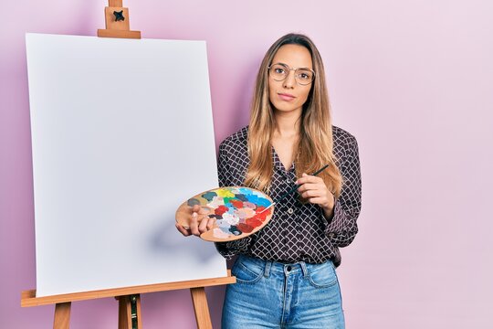 Beautiful hispanic woman standing drawing with palette by painter easel stand relaxed with serious expression on face. simple and natural looking at the camera.