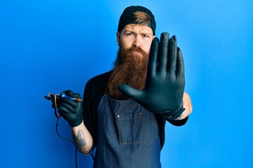 Redhead man with long beard tattoo artist wearing professional uniform and gloves doing stop sing...