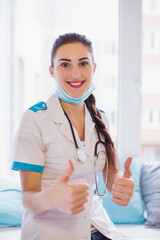 Portrait of the smiling young doctor who is sitting on a sofa in the room and showing thumbs up