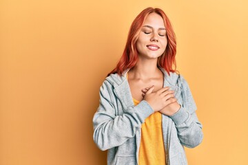 Young beautiful redhead woman wearing casual sporty sweatshirt smiling with hands on chest with closed eyes and grateful gesture on face. health concept.