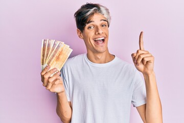 Young hispanic man holding 500 norwegian krone banknotes smiling with an idea or question pointing finger with happy face, number one