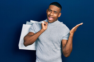 Young black man holding shopping bags celebrating victory with happy smile and winner expression with raised hands
