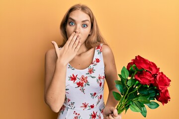 Beautiful blonde caucasian woman holding bouquet of red roses covering mouth with hand, shocked and...