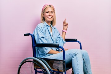 Beautiful blonde woman sitting on wheelchair with a big smile on face, pointing with hand finger to...