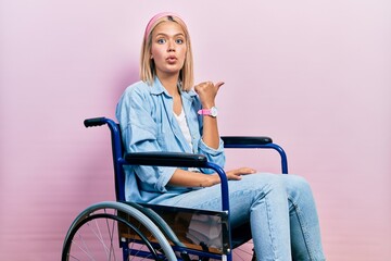 Beautiful blonde woman sitting on wheelchair surprised pointing with hand finger to the side, open...