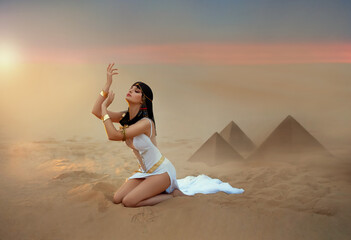 Egypt Style Woman. Sexy beautiful girl goddess Queen Cleopatra sits on sand desert pyramids. Art ancient pharaoh costume white dress gold accessories. Egyptian makeup. hands raised to sky lady praying - obrazy, fototapety, plakaty