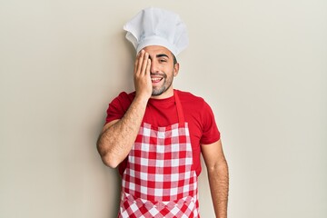 Young hispanic man wearing baker uniform covering one eye with hand, confident smile on face and surprise emotion.