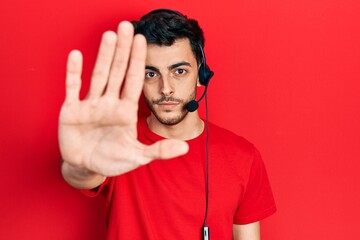 Young hispanic man wearing call center agent headset with open hand doing stop sign with serious and confident expression, defense gesture