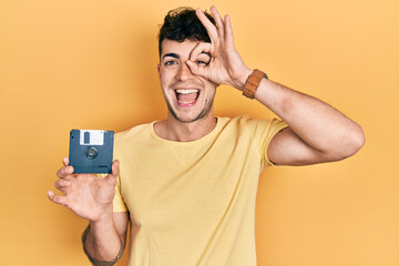 Young hispanic man holding floppy disk smiling happy doing ok sign with hand on eye looking through...