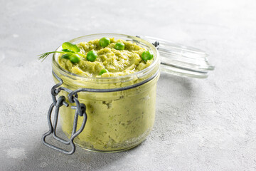 Avocado hummus in a glass jar with chickpeas, green peas, spices on gray background. Healthy vegan...