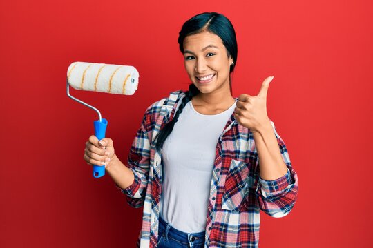 Beautiful hispanic woman holding roller painter smiling happy and positive, thumb up doing excellent and approval sign