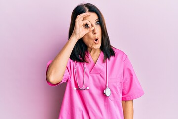 Middle age brunette woman wearing doctor uniform and stethoscope doing ok gesture shocked with surprised face, eye looking through fingers. unbelieving expression.