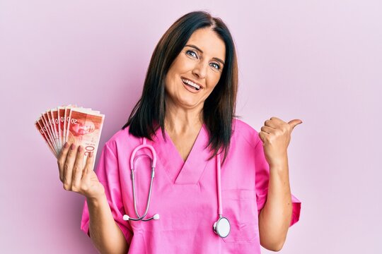 Middle age brunette doctor woman holding 20 shekels banknotes pointing thumb up to the side smiling happy with open mouth