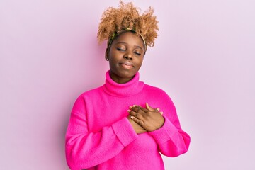 Young african woman with afro hair wearing casual winter sweater smiling with hands on chest, eyes closed with grateful gesture on face. health concept.