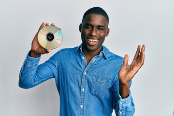 Young african american man holding compact disc celebrating victory with happy smile and winner...