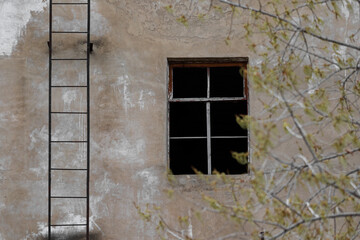 A broken window and a fire escape in an old abandoned building with a poorly painted beige facade and a vintage-looking wooden window frame with branches of wood in the foreground is white plaster.