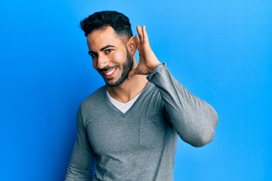 Young hispanic man wearing casual white t shirt smiling with hand over ear listening and hearing to rumor or gossip. deafness concept.