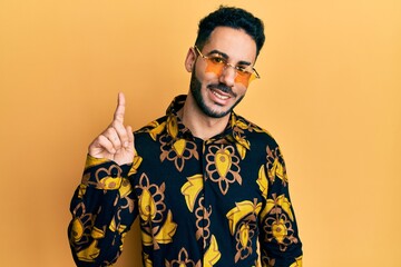 Young hispanic man wearing stylish sunglasses smiling with an idea or question pointing finger up with happy face, number one