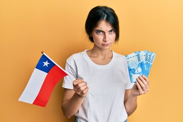 Young caucasian woman holding chile flag and chilean pesos banknotes skeptic and nervous, frowning upset because of problem. negative person.