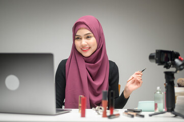 young muslim woman entrepreneur working with laptop presents cosmetic products during online live stream over white background studio, selling online and beauty blogger concept