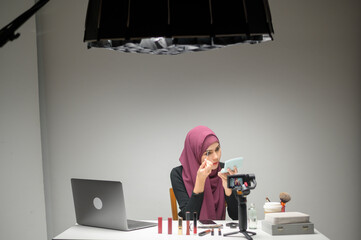 Plakat young muslim woman entrepreneur working with laptop presents cosmetic products during online live stream over white background studio, selling online and beauty blogger concept