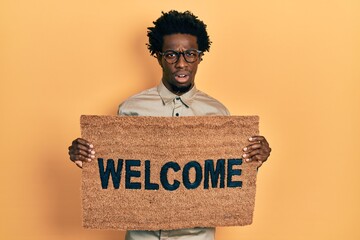 Young african american man holding welcome doormat in shock face, looking skeptical and sarcastic,...