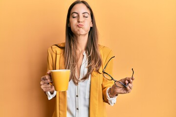 Young beautiful woman wearing business style drinking cup of coffee puffing cheeks with funny face....