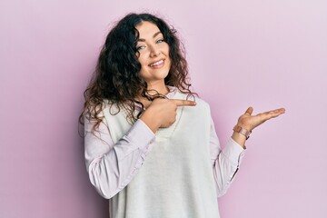 Young brunette woman with curly hair wearing casual clothes amazed and smiling to the camera while presenting with hand and pointing with finger.