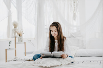 brunette preteen girl in jeans and a white shirt is resting and reading a book on a bed in a bright room with a Balinese-style interior