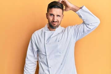 Handsome man with beard wearing professional cook uniform confuse and wonder about question. uncertain with doubt, thinking with hand on head. pensive concept.