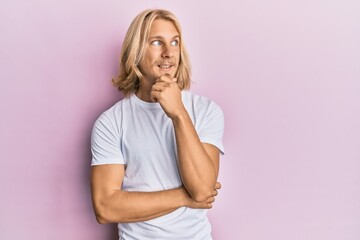 Caucasian young man with long hair wearing casual white t shirt with hand on chin thinking about...