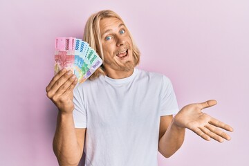 Caucasian young man with long hair holding indonesian rupiah banknotes celebrating achievement with happy smile and winner expression with raised hand