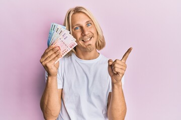 Caucasian young man with long hair holding new taiwan dollars banknotes smiling happy pointing with hand and finger to the side