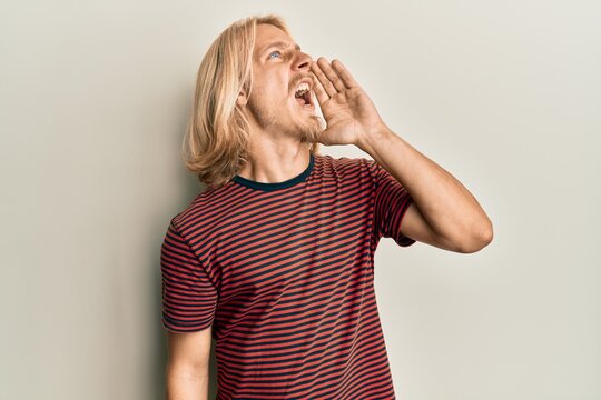 Caucasian young man with long hair wearing casual striped t shirt shouting and screaming loud to side with hand on mouth. communication concept.