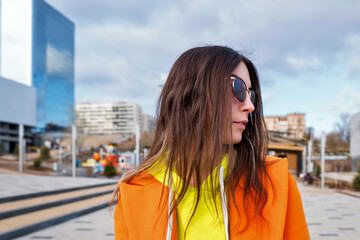Fototapeta na wymiar Close up portrait Millennial Hipster Girl in lilac sunglasses, orange coat, yellow hoodie posing at urban background. Carefree good-looking young woman walks city street.