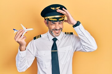Handsome middle age man with grey hair wearing airplane pilot uniform holding toy plane stressed...