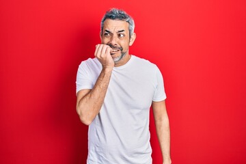 Handsome middle age man with grey hair wearing casual white t shirt looking stressed and nervous with hands on mouth biting nails. anxiety problem.