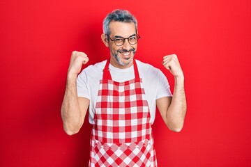 Naklejka premium Handsome middle age man with grey hair wearing apron screaming proud, celebrating victory and success very excited with raised arms