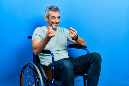Handsome middle age man with grey hair sitting on wheelchair doing money gesture with hands, asking for salary payment, millionaire business