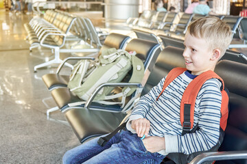 Happy junior schoolboy with orange backpack sits waiting for flight on comfortable chair in...