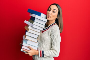 Young brunette student girl holding a pile of books making fish face with mouth and squinting eyes, crazy and comical.