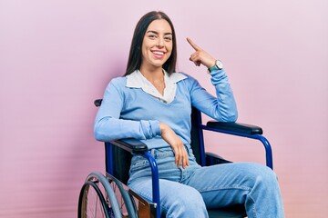 Beautiful woman with blue eyes sitting on wheelchair smiling pointing to head with one finger,...