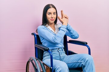 Beautiful woman with blue eyes sitting on wheelchair holding symbolic gun with hand gesture,...