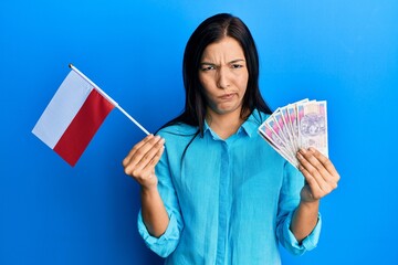 Young latin woman holding poland flag and zloty banknotes skeptic and nervous, frowning upset because of problem. negative person.
