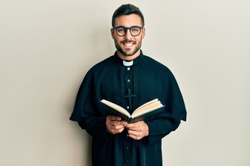 Young hispanic priest man holding bible smiling with a happy and cool smile on face. showing teeth.