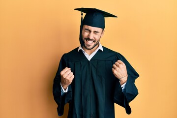 Young hispanic man wearing graduation cap and ceremony robe excited for success with arms raised...