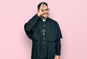 Young hispanic man wearing priest uniform smiling happy doing ok sign with hand on eye looking through fingers